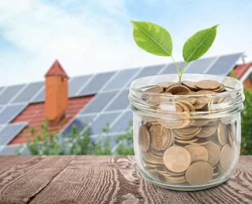 Glass jar with coins and plant against house with installed solar panels on roof. Economic benefits of renewable energy