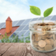 Glass jar with coins and plant against house with installed solar panels on roof. Economic benefits of renewable energy