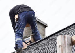 Roofer construction worker repairing flashing around chimney on grey slate shingles roof of domestic house, sky background with copy space.