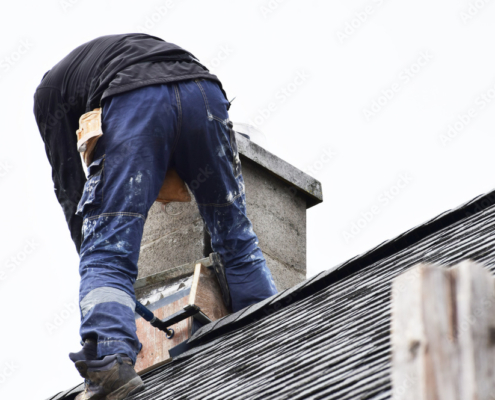 Roofer construction worker repairing flashing around chimney on grey slate shingles roof of domestic house, sky background with copy space.