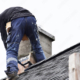 Roofer construction worker repairing flashing around chimney on grey slate shingles roof of domestic house, sky background with copy space.