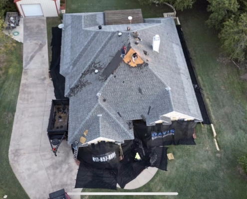 Aerial of a sprawling home being reroofed, crew working the ridge with a loaded debris trailer in the drive.