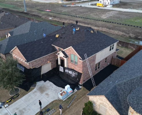 Aerial of a two-story brick home reroof with crew tearing off shingles and new bundles staged in the drive.