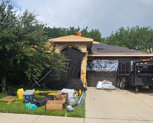 Brick home mid re-roof with felt-covered sections, a dump trailer, and a roofer working up top.