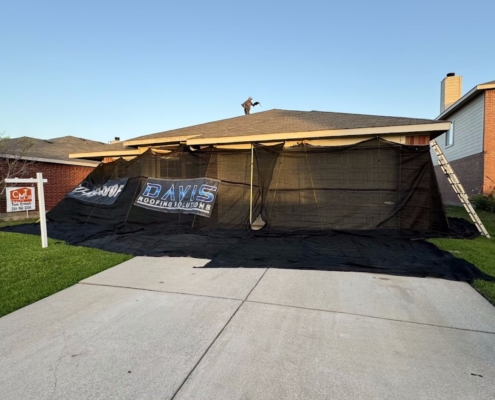 Single-story home enclosed in black ground-protection netting as a roofer works on the roof at sunset.