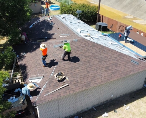 Overhead view of roofers working across a brown shingle roof with a loaded dump trailer below