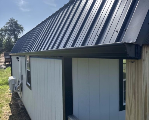 Close-up of a dark standing-seam metal roof and drip edge along the eave of a white cabin.