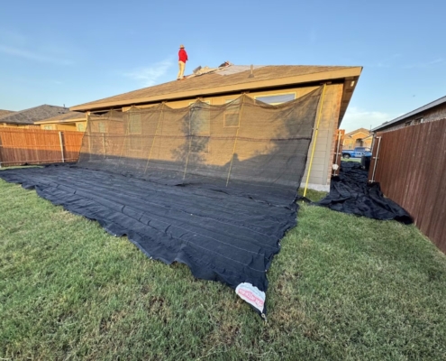 Home surrounded by black debris-catch netting as a roofer works on the roof in evening light.
