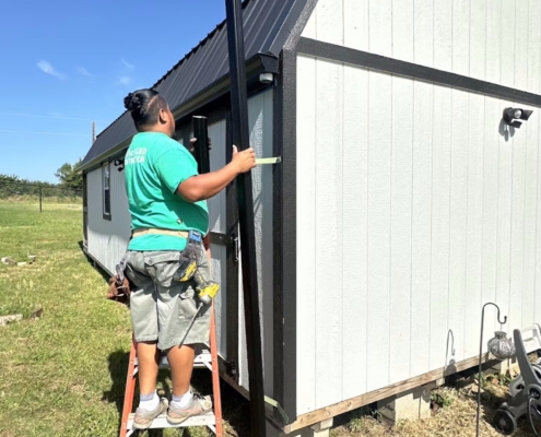 Worker on a ladder fastening a black downspout to a white cabin with a dark metal roof.