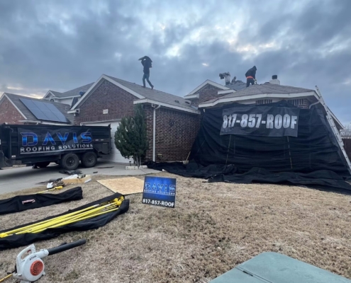 Roofers tearing off homes at dusk with a Davis Roofing dump trailer and job banners on site