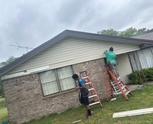 Two roofers on ladders replacing fascia and soffit along a single-story brick home's eave.