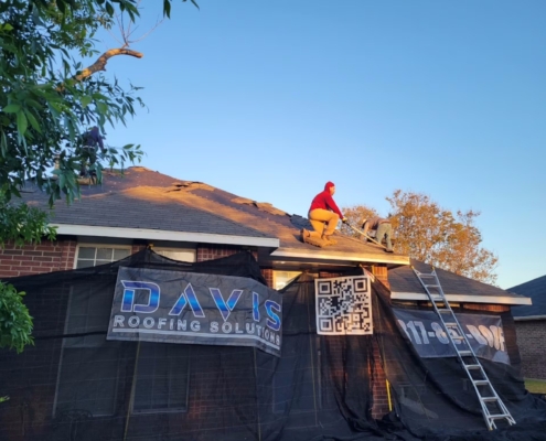 Roofer tearing off old shingles at golden hour, exposing decking above the job-site protection.