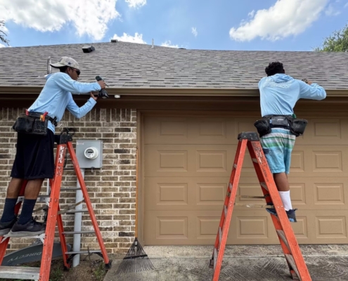 Two roofers on ladders drilling gutters and fascia along the edge of a brick home's roof.