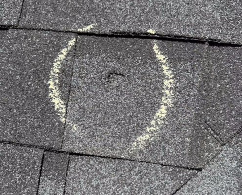Storm-damaged asphalt shingle with a hail impact mark circled in chalk during a roof inspection.