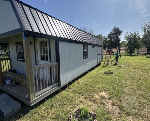 New black-trimmed metal roof on a white lofted cabin with a covered porch in a rural yard.
