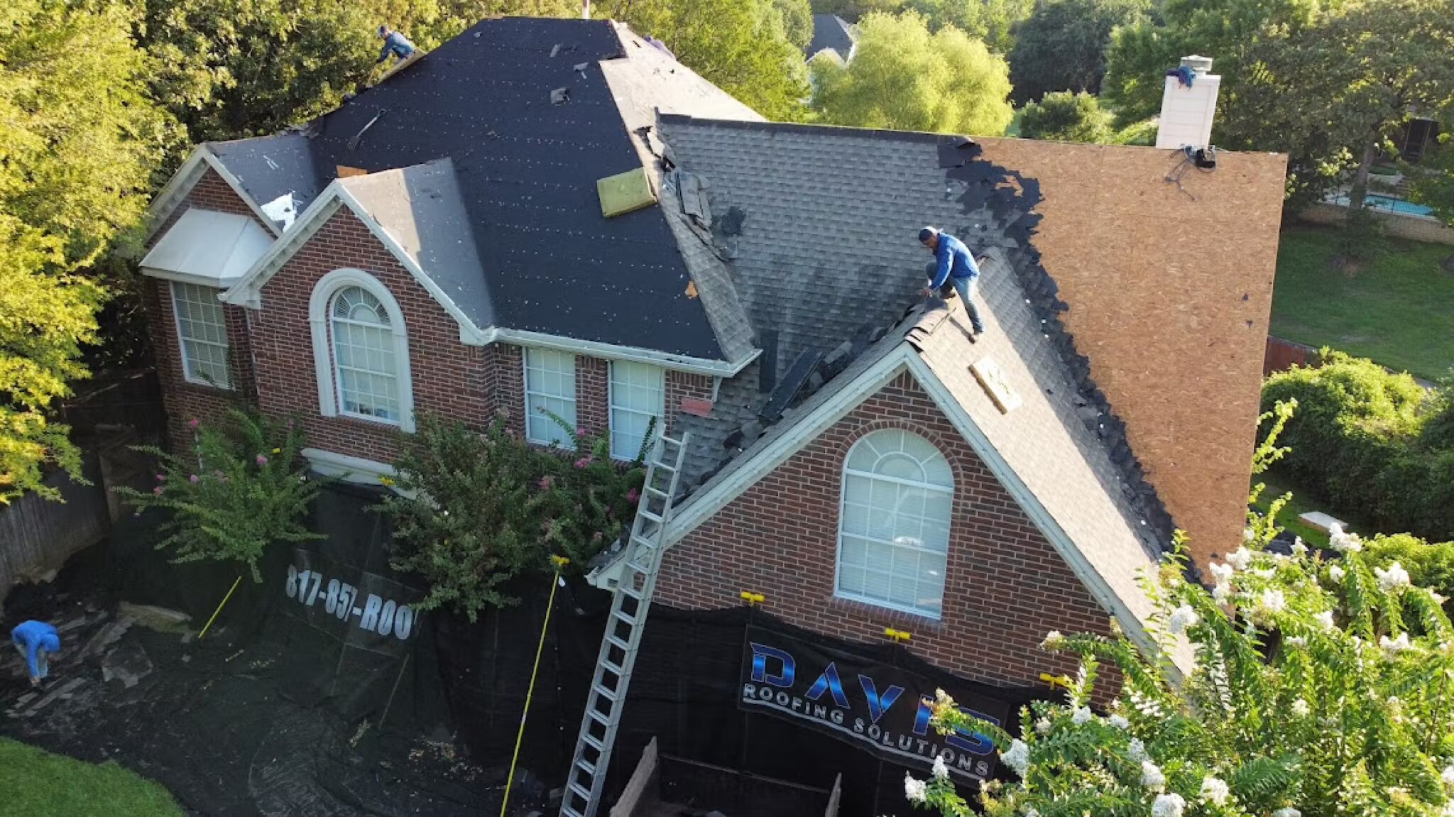 Aerial of a worker stripping old shingles on a brick home with a Davis Roofing banner hung below