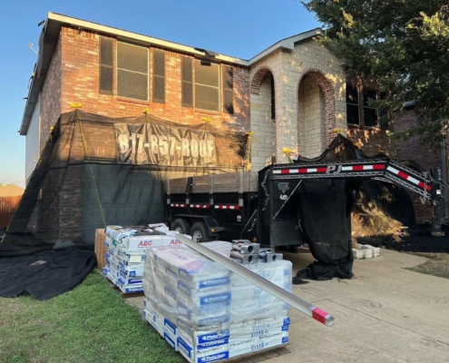Pallets of shingles and a dump trailer staged at a two-story home during a roof replacement