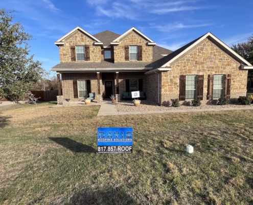 Two-story stone and brick home with a freshly installed shingle roof under a clear blue Texas sky.