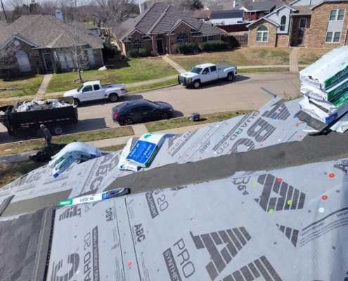 Roof deck covered in synthetic underlayment with shingle bundles staged and a debris trailer at the curb.