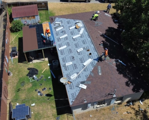 Aerial of roofers installing underlayment on a home roof with a dump trailer parked alongside