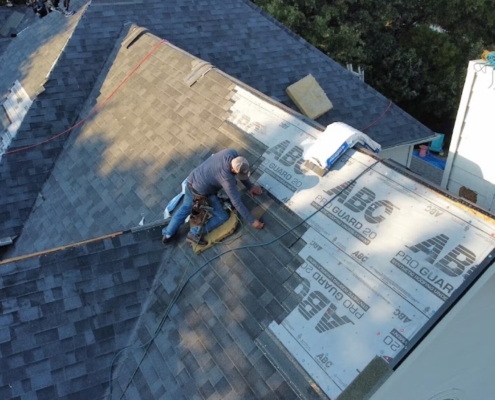 Roofer kneeling to install synthetic underlayment along a roof valley near a chimney