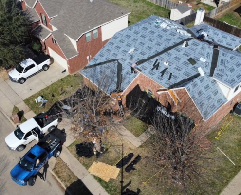 Drone view of a brick home with synthetic underlayment down and work trucks parked at the curb